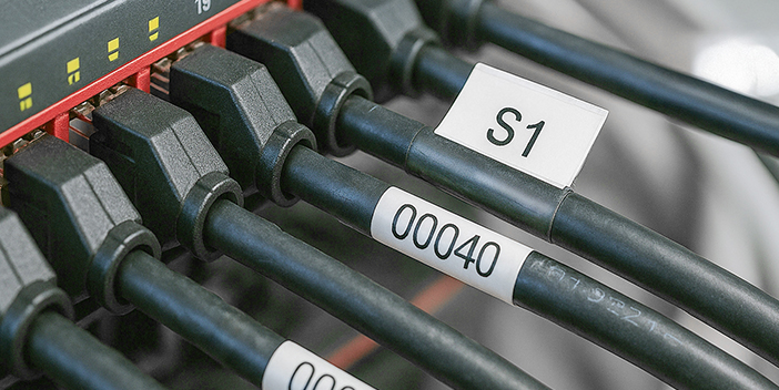Nahaufnahme von mehreren schwarzen Kabeln, die an ein Panel angeschlossen sind, Teil eines Schaltschrankes. Jedes Kabel ist mit einem wei&szlig;en Etikett versehen, das alphanumerische Codes wie &bdquo;S1&ldquo; und &bdquo;00040&ldquo; anzeigt.