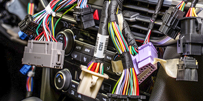 Nahaufnahme von mehreren Kabelb&auml;umen im Armaturenbrett eines Fahrzeugs, die mit verschiedenen Steckverbindern verbunden sind. Ein wei&szlig;er Kabelmarkierer mit gedrucktem Text, Barcode und Seriennummer ist an einem der Kabelb&uuml;ndel angebracht.