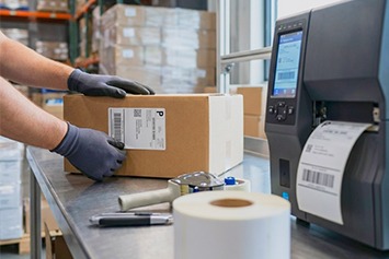 Eine Person bringt manuell ein Versandetikett auf einen Karton am Packtisch in der Versandlogistik auf.
