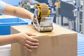 Ein Karton wird im Verpackungsprozess manuell mit Paketband und einem Handabroller verschlossen.
